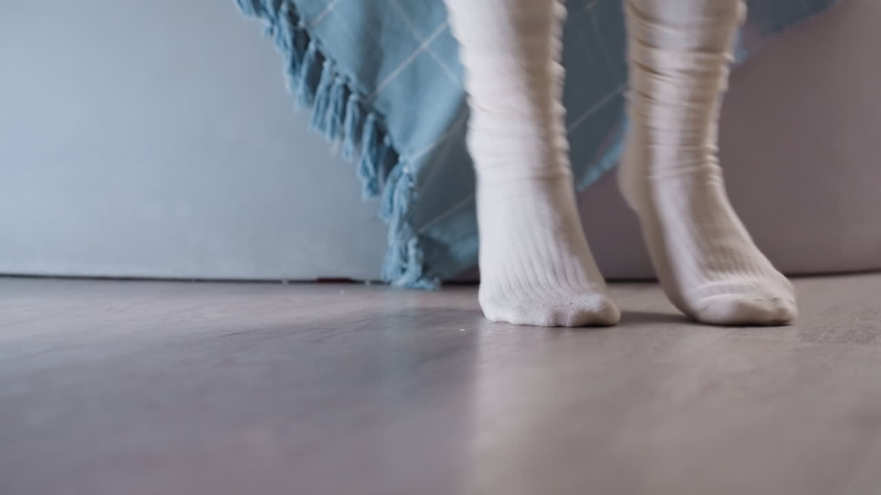 Close view of person in white socks and gray leggings jumping on wooden floor near bed with blue blanket, capturing cozy lifestyle, relaxation, casual home atmosphere and everyday comfort indoors