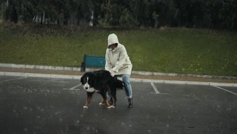 Mujer-Feliz-Con-Una-Chaqueta-Blanca-Acariciando-A-Su-Perro-Grande-Mientras-Camina-Bajo-Una-Fuerte-Lluvia-En-El-Parque