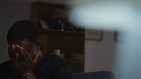 unhappy and depressed young man sitting on sofa at home at night shaking head