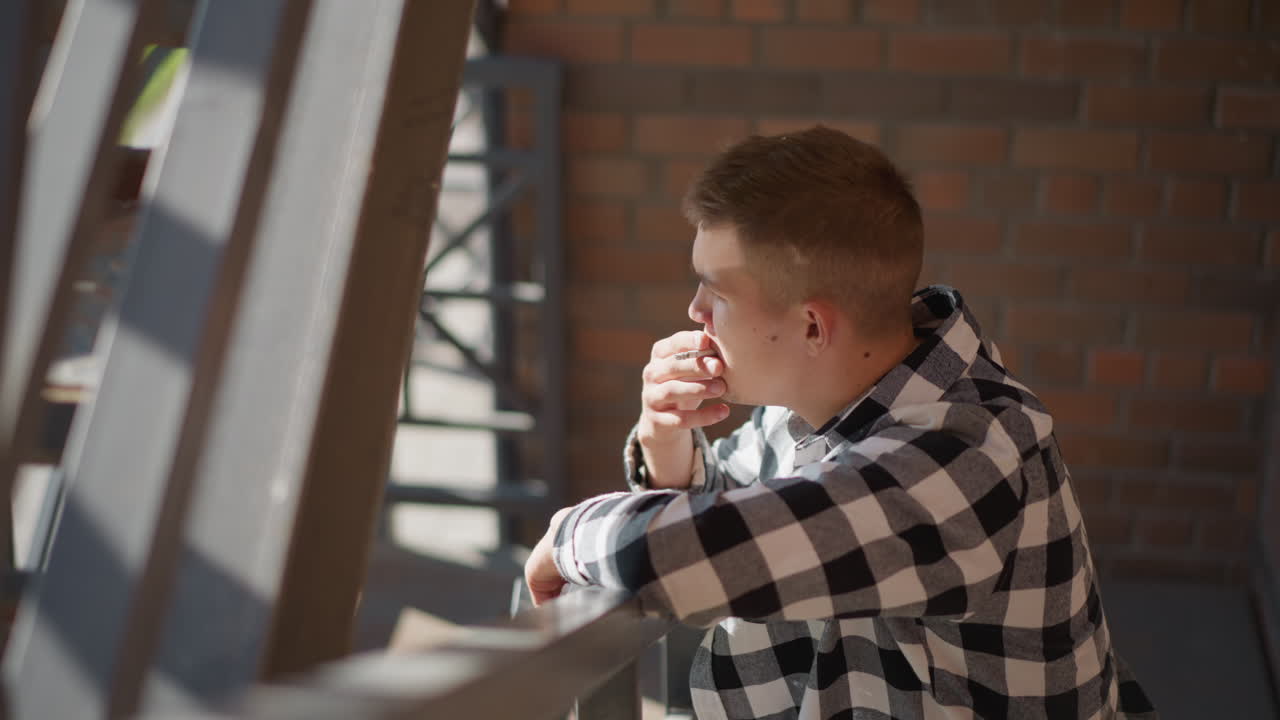 man in checkered shirt rests on iron railing smoking cigarette while holding pack in hand bright sunlight filters through metal stair casting patterned shadows across outdoor brick wall
