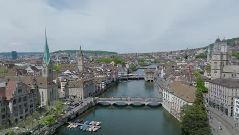 Fast-forward-moving,-descending-drone-shot-of-Zurich-city,-showing-old-town-architecture,-clock-towers-and-cities-skyline-with-river-in-the-foreground