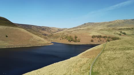 Aerial-shot-of-Kinder-Reservoir-with-Geese-on-the-water-and-people-in-the-distance-walking-on-the-other-side-and-the-valley-climb-up-the-Kinder-Scout-Peak,-Peak-District,-UK
