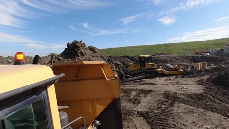 Toma-De-Cámara-De-Acción-De-Un-Camión-Volquete-Volcando-Una-Carga-De-Tierra-Frente-A-Una-Excavadora-Y-Un-Rodillo