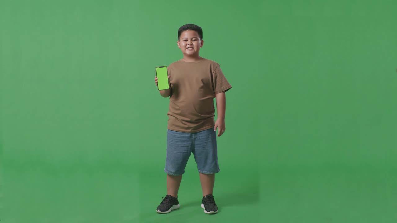 cuerpo lleno de niño asiático con una sonrisa que muestra pantalla verde teléfono móvil mientras está de pie en la pantalla verde