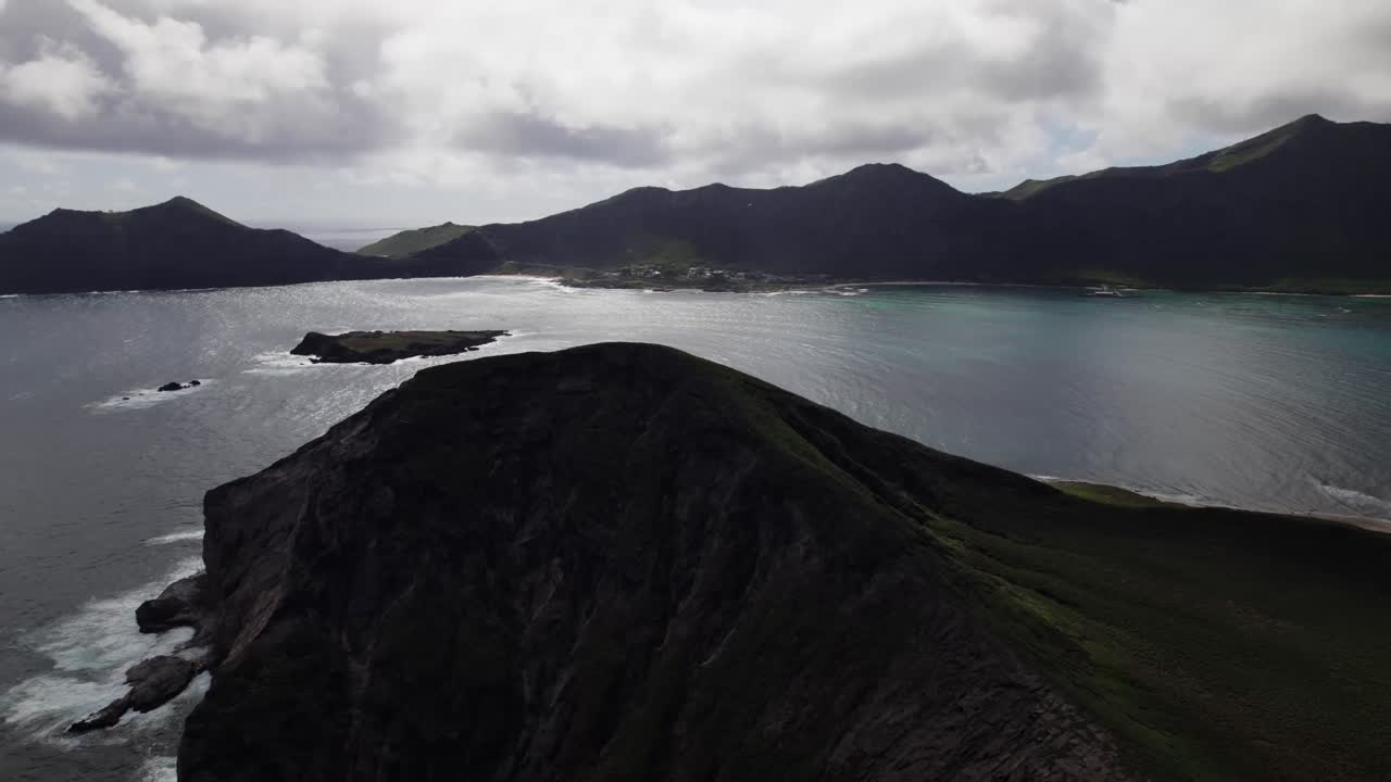 Drone shot of Makapuʻu's rugged cliff edge overlooking the turquoise waters of Waimānalo Bay, backed by volcanic ridges and dramatic skies.