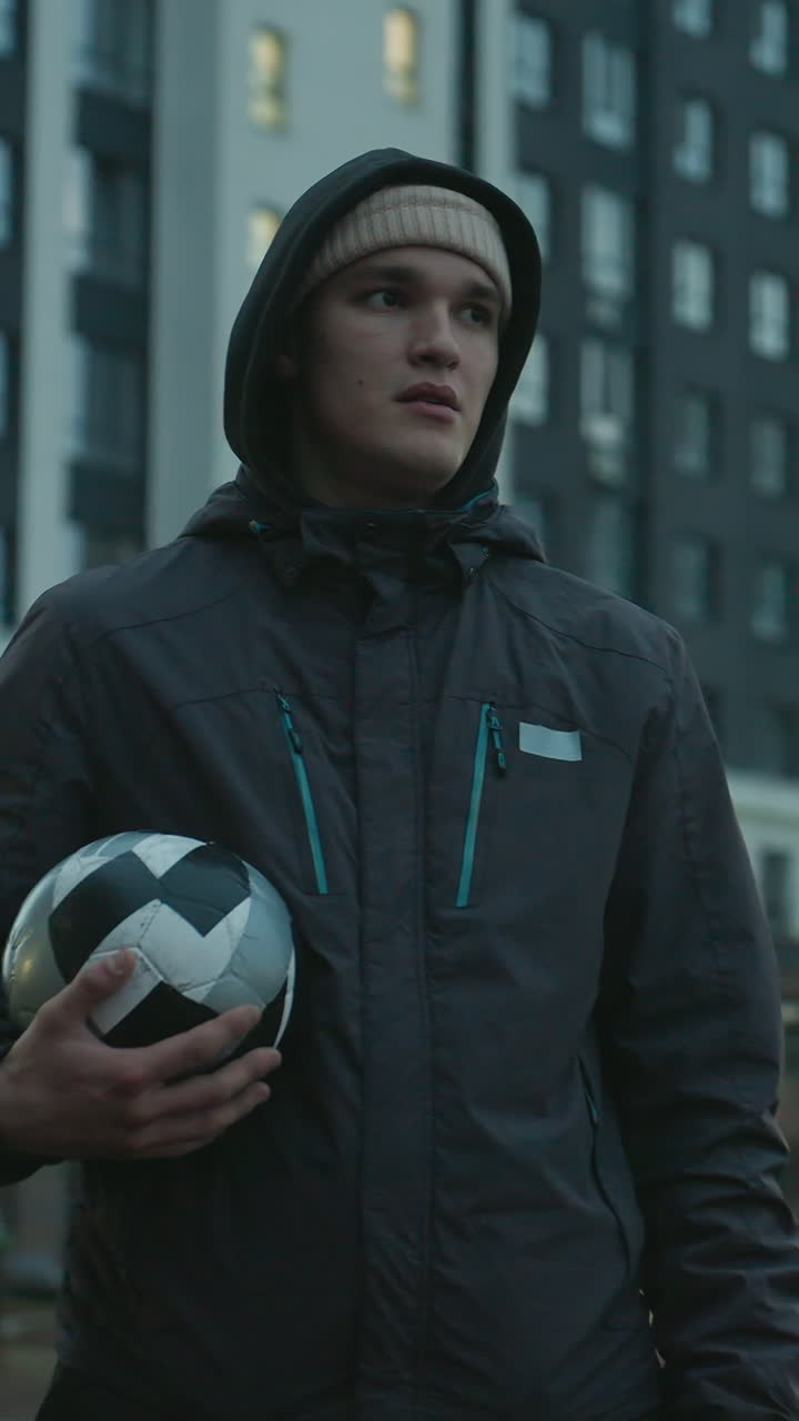 Man holding soccer ball standing confidently in modern sport arena surrounded by tall residential buildings under a cloudy evening sky