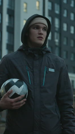 man holding soccer ball standing confidently in modern sport arena surrounded by tall residential buildings under a cloudy evening sky