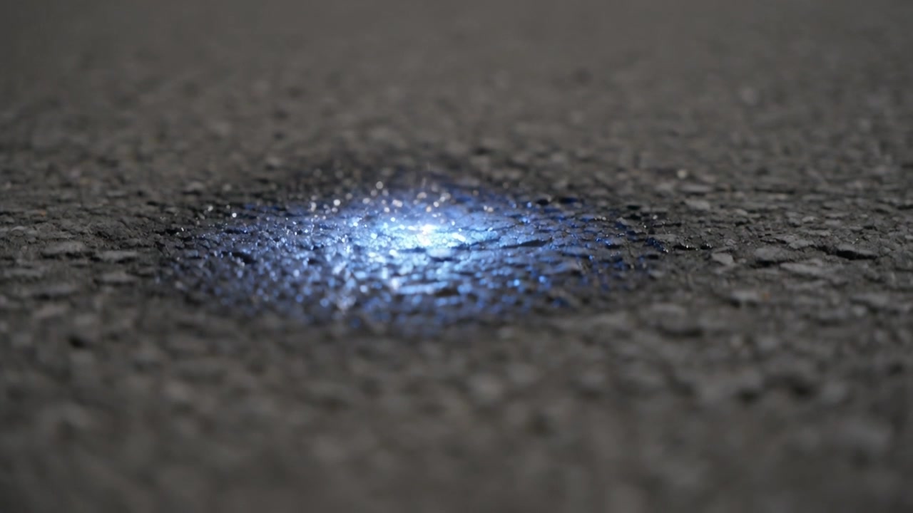 Appearing bright blue point expanding into shallow glowing patch on rough asphalt, sparks subsiding