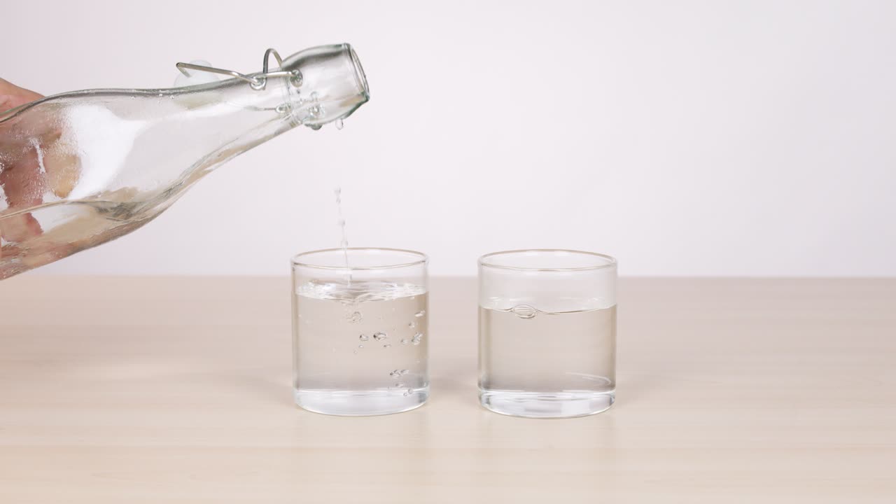 Hands pour water from glass bottle into two clear glasses, bright lighting, static camera