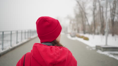 vista posterior de una mujer caminando por un camino entrelazado cerca de una barandilla de hierro con gorra roja y chaqueta con auriculares alrededor del cuello en un parque de invierno nevado con árboles borrosos y postes de lámpara en el fondo