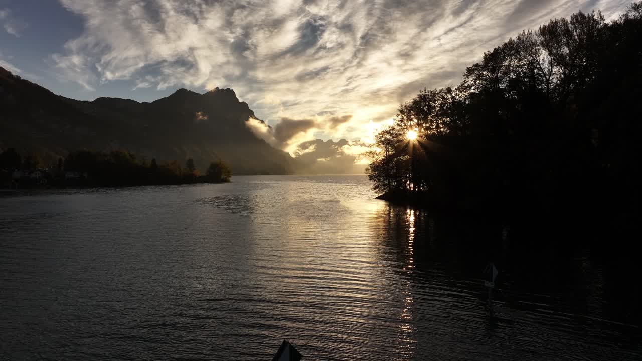 Serene atmosphere at Switzerland's Lake Walensee, with low-angle sunlight reflecting on calm waters and a lone tree gracing the shore. A tranquil scene capturing nature's quiet beauty.