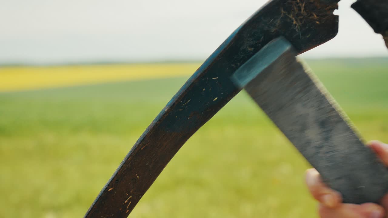 Man sharpening a scythe blade by hand in a rural field. Traditional farming activity captured in close-up with natural light and scenic countryside background. Close Up.