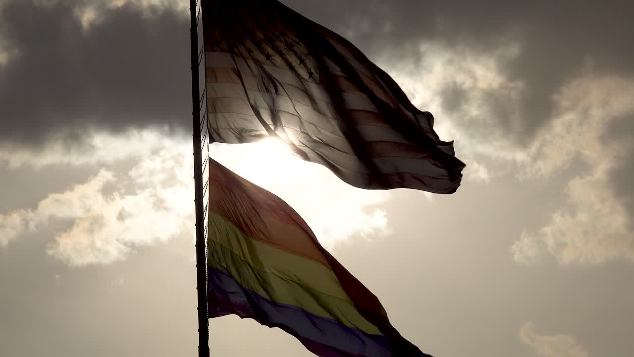Rainbow LGBT gay pride flag waving in the wind 4 and the American flag waving with sun backlit under gray clouds 4k