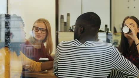 la cámara se enfoca en un grupo multiétnico de amigos a través de la ventana hablando y bebiendo café sentados en una mesa en un café