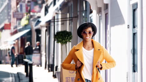 Una-Joven-Negra-De-Moda-Con-Jeans-Azules-Y-Un-Abrigo-De-Guisante-Amarillo-Desabrochado-Caminando-Por-Una-Calle-Pasando-Por-Tiendas-En-Un-Día-Soleado,-Sonriendo,-De-Cerca