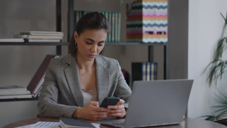 Indian-hispanic-woman-spend-free-time-using-internet-social-media-network-website-read-multimedia-news.-Business-arabian-lady-is-chatting-on-mobile-phone-and-working-with-laptop-indoors-in-apartment.