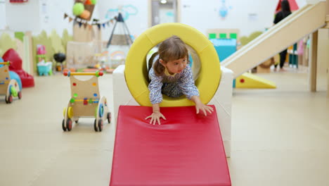 happy little girl crawls through pipe and slides down at playroom