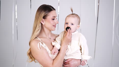 Long-haired blonde woman in white dress holding her baby in hands. Mother gives a pacifier to her kid. Studio footage.