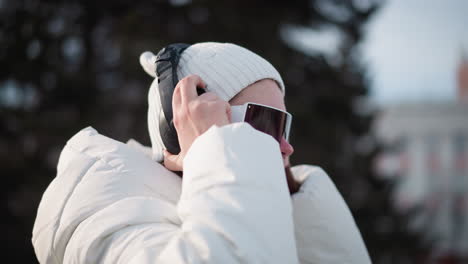 side view of young woman wearing wired ear piece nodding head under sunny winter sky while adjusting headphone headset over beanie and goggles on snow dusted plaza with blurred traffic backdrop