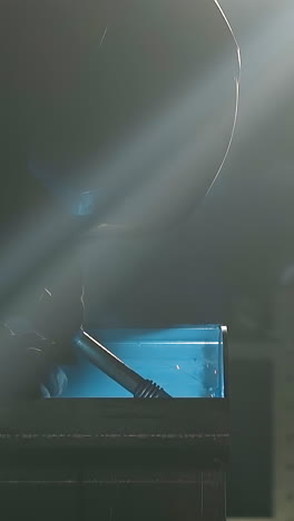silhouette of employee connecting steel details with welding machine in workshop closeup. smoke and flying sparks in dark factory enclosure lit by sunbeam