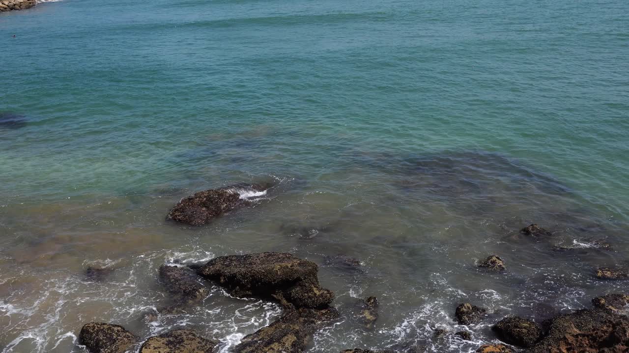 Onde delicate si infrangono sugli scogli lungo la riva, con una lunga distesa di acqua turchese sullo sfondo, creando una scena davvero incantevole