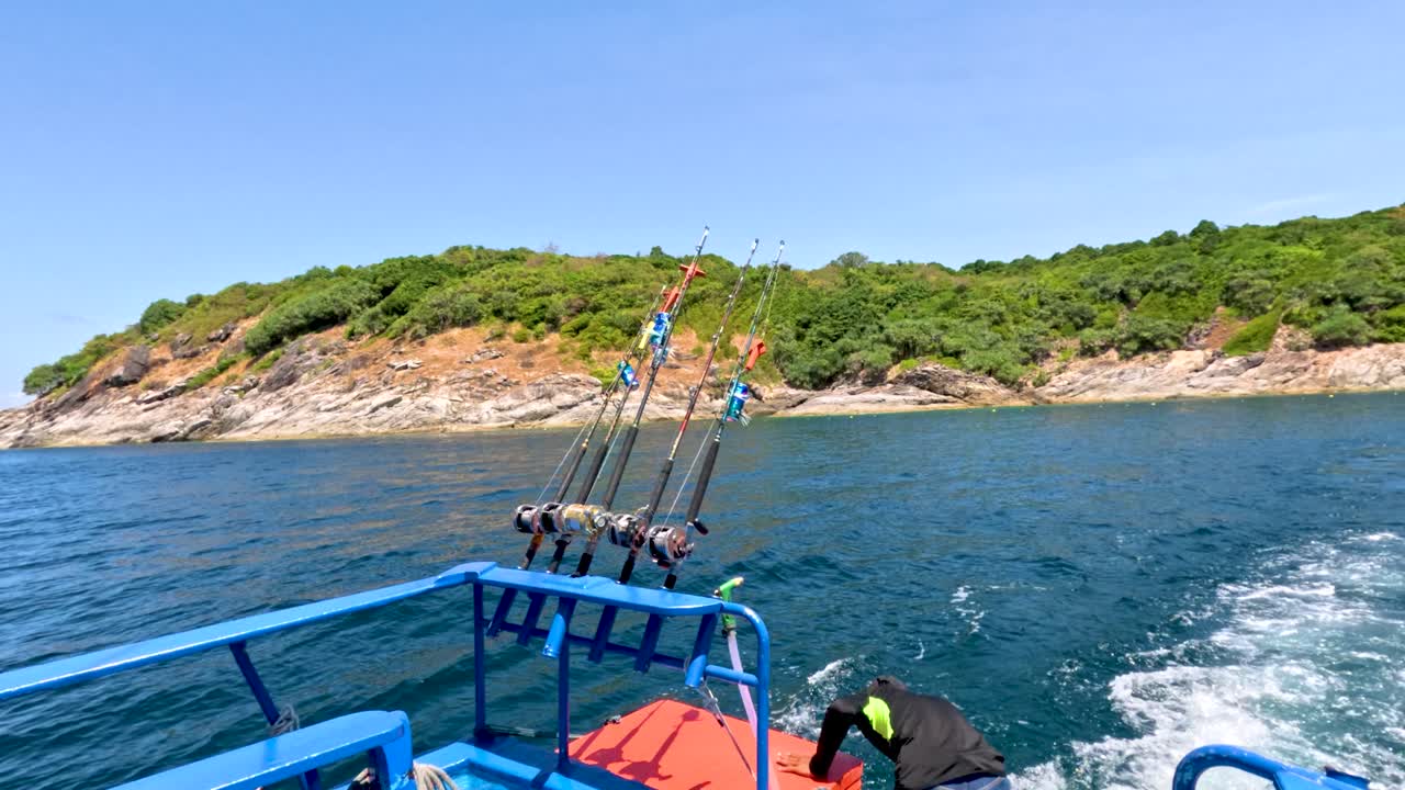 Man adjusts fishing rods on moving boat under bright daylight near lush Phuket shoreline