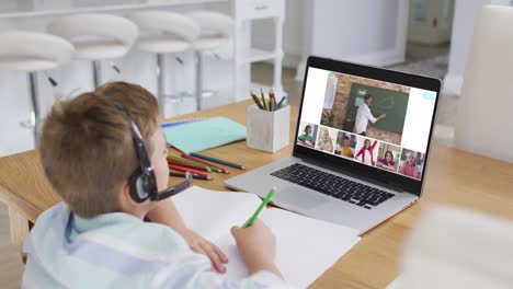 Schoolboy-using-laptop-for-online-lesson-at-home,-with-diverse-teacher-and-class-on-screen