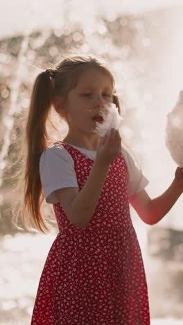 little children tear and eat pieces of cotton candy standing against fountain in park slow motion. traditional dessert in urban garden. happy kids
