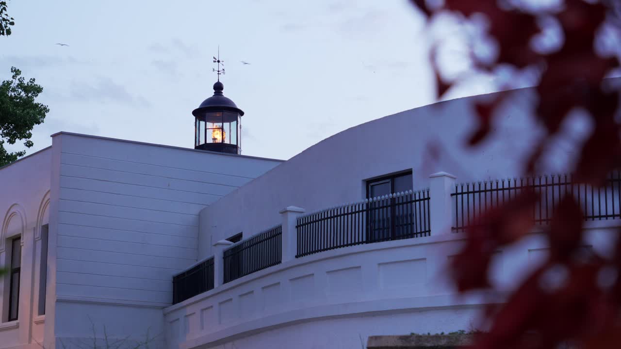 View of a small modern lighthouse atop a white building glowing at dusk, with soft evening sky and foliage in foreground. Filmed in Constanța, Romania