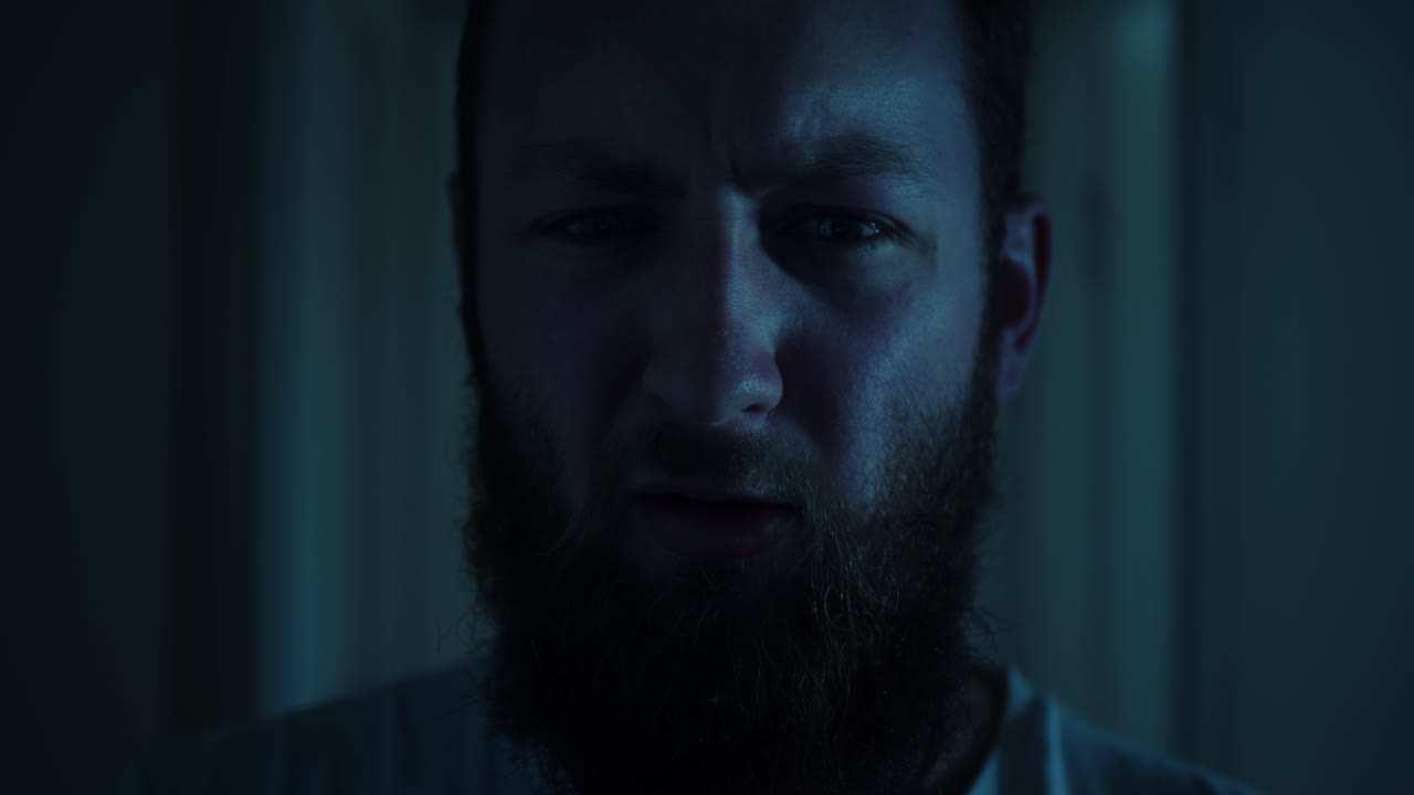 un hombre con barba se ve de cerca, su rostro parcialmente iluminado por la tenue luz de la luna azulada. la expresión es tensa, como si estuviera profundamente en pensamiento o descubriendo algo inquietante antes de caminar hacia adelante.