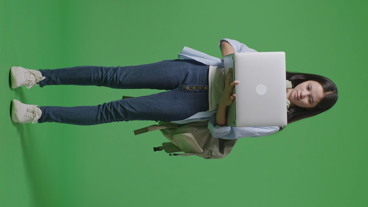 cuerpo lleno de estudiante adolescente asiática con una mochila sonriendo y escribiendo en una computadora portátil mientras está de pie en el estudio de fondo de la pantalla verde