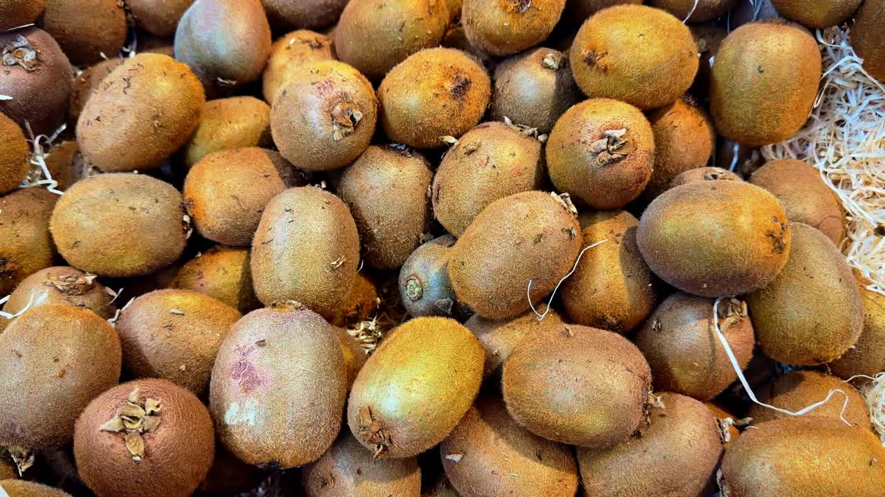 Ripresa dall'alto con movimento panoramico che mostra kiwi dorati maturi presentati in un autentico mercato contadino, evidenziando la buccia marrone pelosa e le imperfezioni naturali