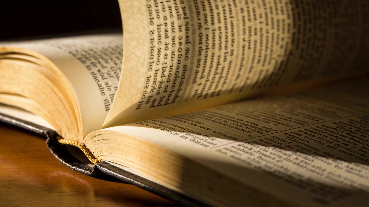 Sollevando e girando la pagina di un libro rilegato aperto a causa di una corrente d'aria sulla scrivania, rivelando il testo sotto una lampada calda.