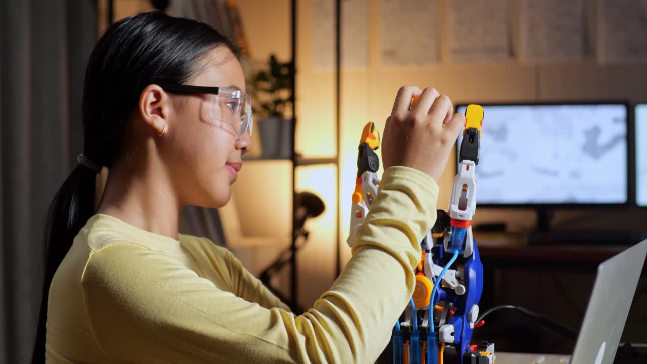 un primer plano de una adolescente asiática sonriente con una mano de cyborg mostrando los pulgares hacia arriba gestando hacia la cámara mientras trabaja en una computadora portátil en casa