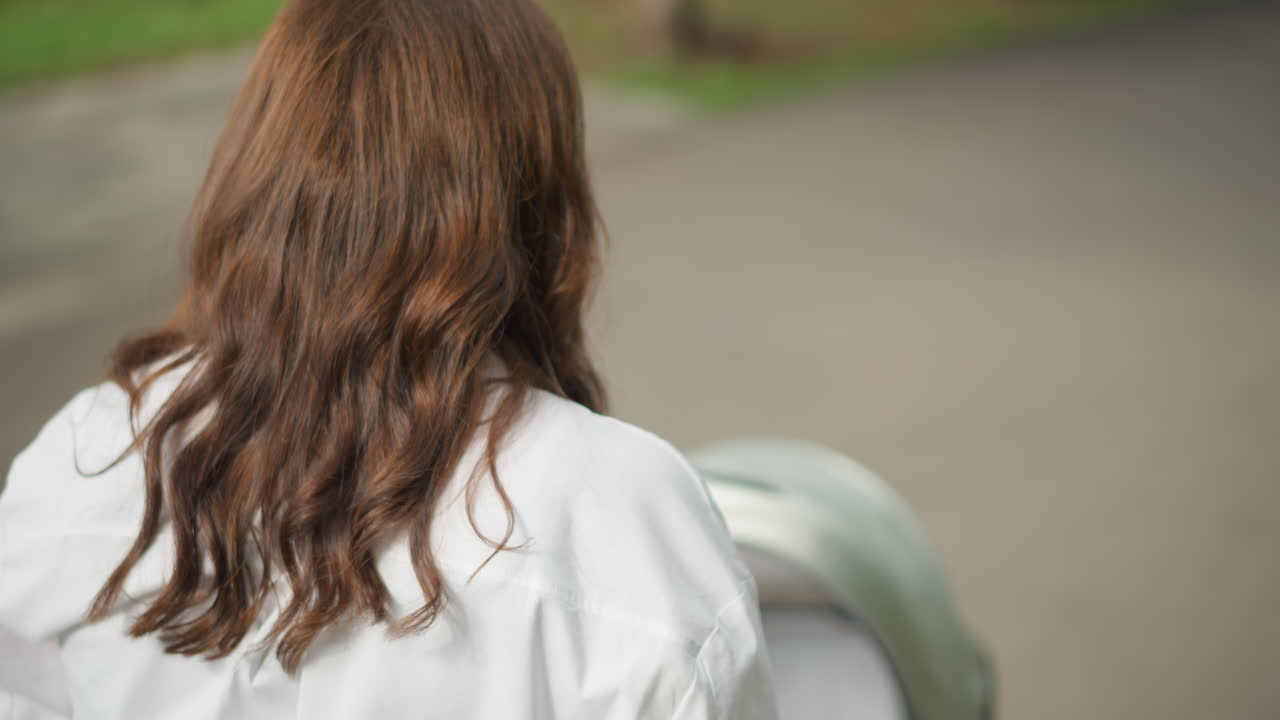 小道をベビーカーを押す女性の後ろ姿。波打つ茶色の髪と白いシャツが特徴的で、物思いにふけるような都会の朝の風景。柔らかい日差しの中、ゆっくりとしたペースで歩き、公園の木々と舗装された道が続く。親としての穏やかな様子が伝わる。