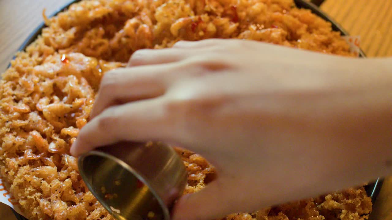 Hand pours chili sauce over golden fried shrimp pancake, top-down view, natural indoor lighting