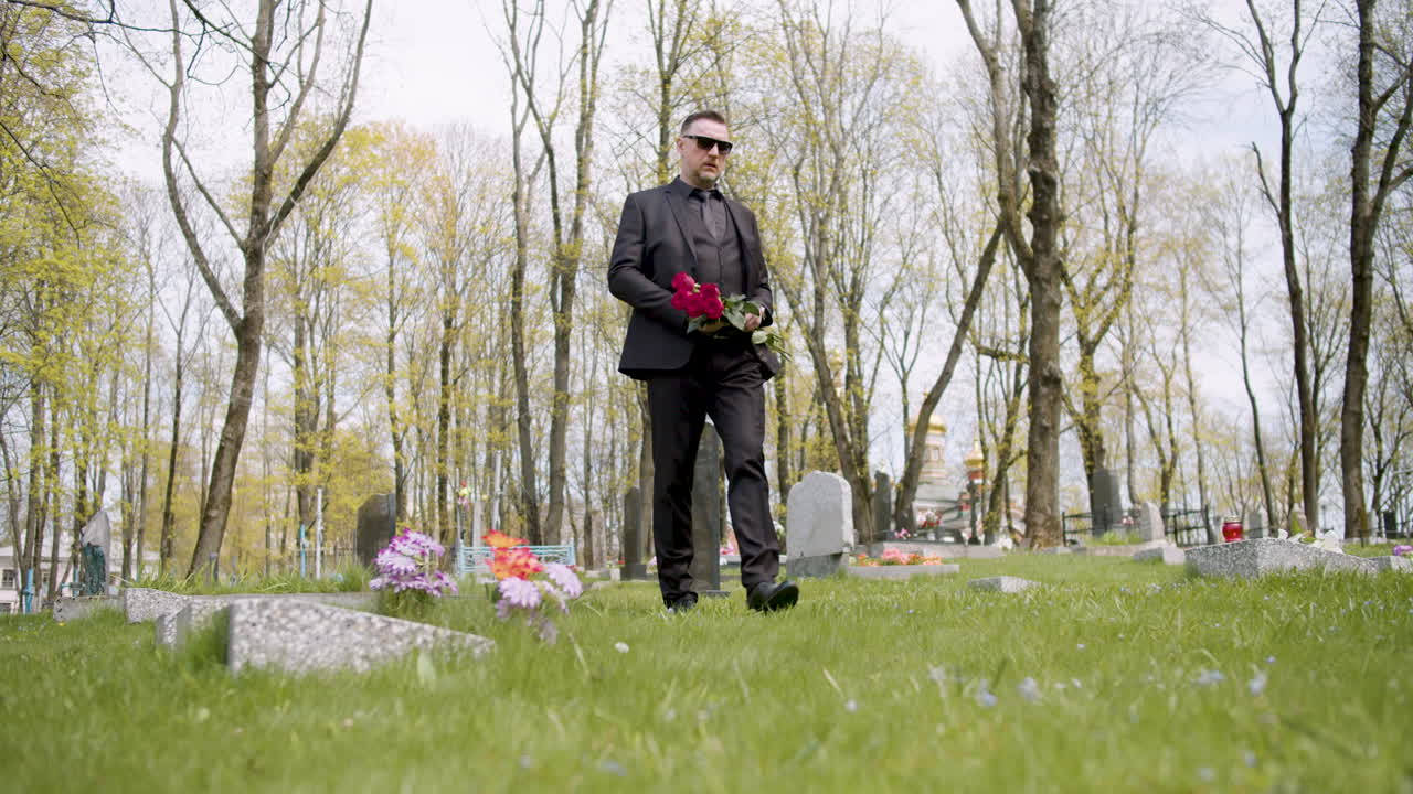 빨간 장미를 들고 묘지 2에서 걷는 검은 양복에 슬픈 남자