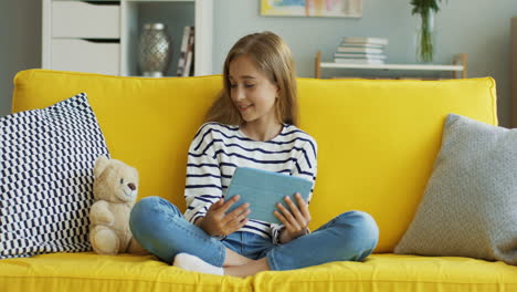 Adolescente-Rubia-Sentada-En-Un-Sofá-Amarillo-En-Casa-Y-Viendo-Algo-En-La-Tableta,-Luego-Mira-Y-Sonríe-A-La-Cámara