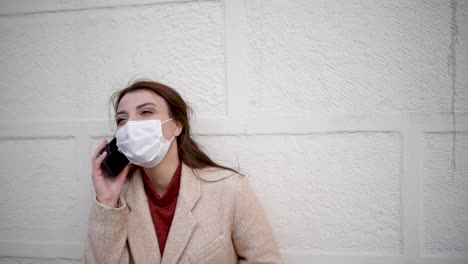 Beautiful-girl-wearing-protective-medical-mask-and-fashionable-clothes-speaks-with-a-smart-phone-in-front-of-wall