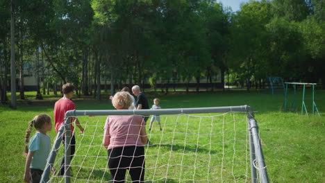 family enjoys an outdoor soccer game on a sunny day as the son in white kicks the ball into the net, with their mom guarding the goal post and siblings eagerly watching and playing along