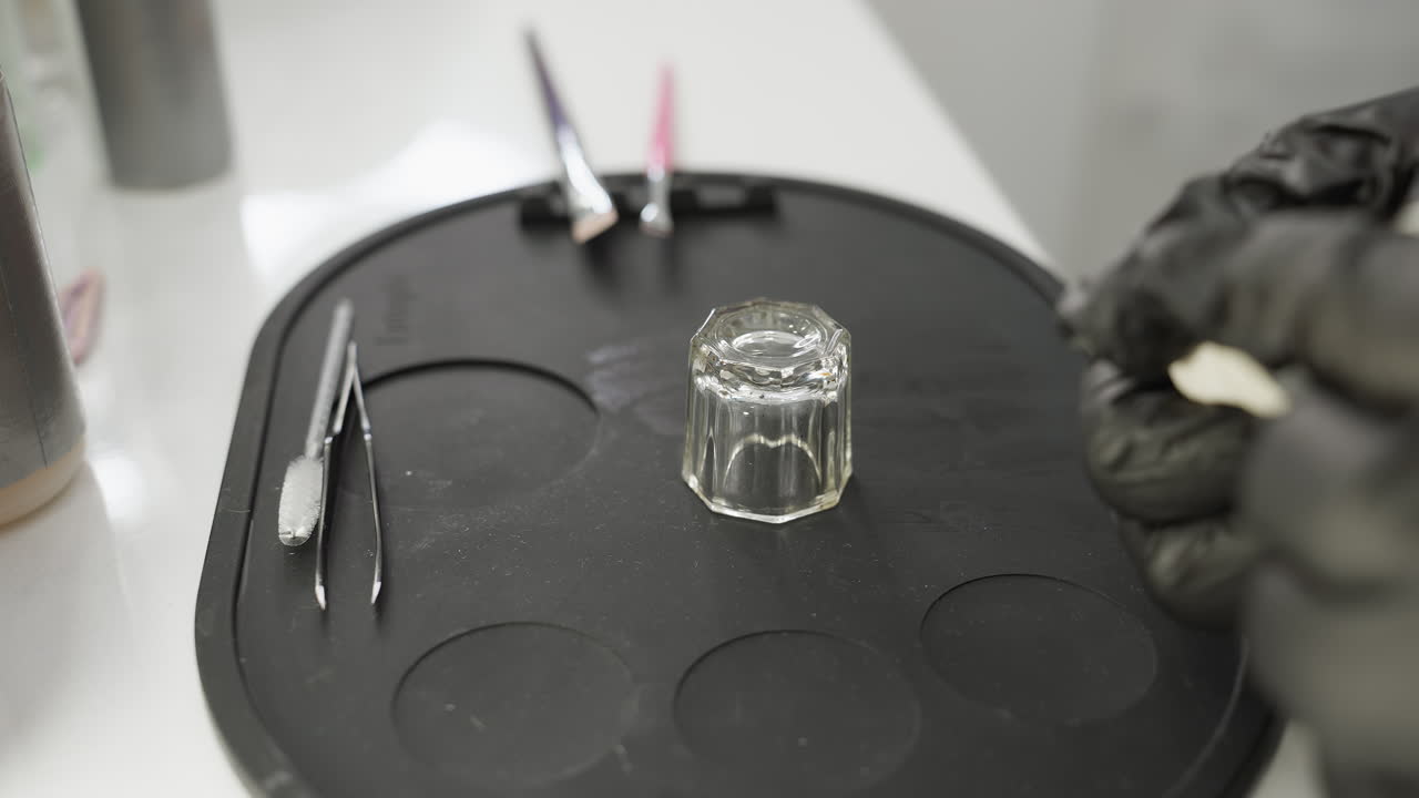 Beautician wearing black gloves prepares for eyebrow session, reaching toward tools on black silicone mat with glass container, tweezers, brushes, and other small instruments neatly arranged