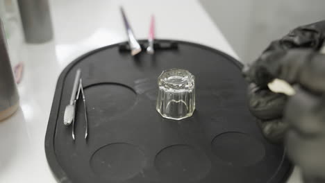 beautician wearing black gloves prepares for eyebrow session, reaching toward tools on black silicone mat with glass container, tweezers, brushes, and other small instruments neatly arranged