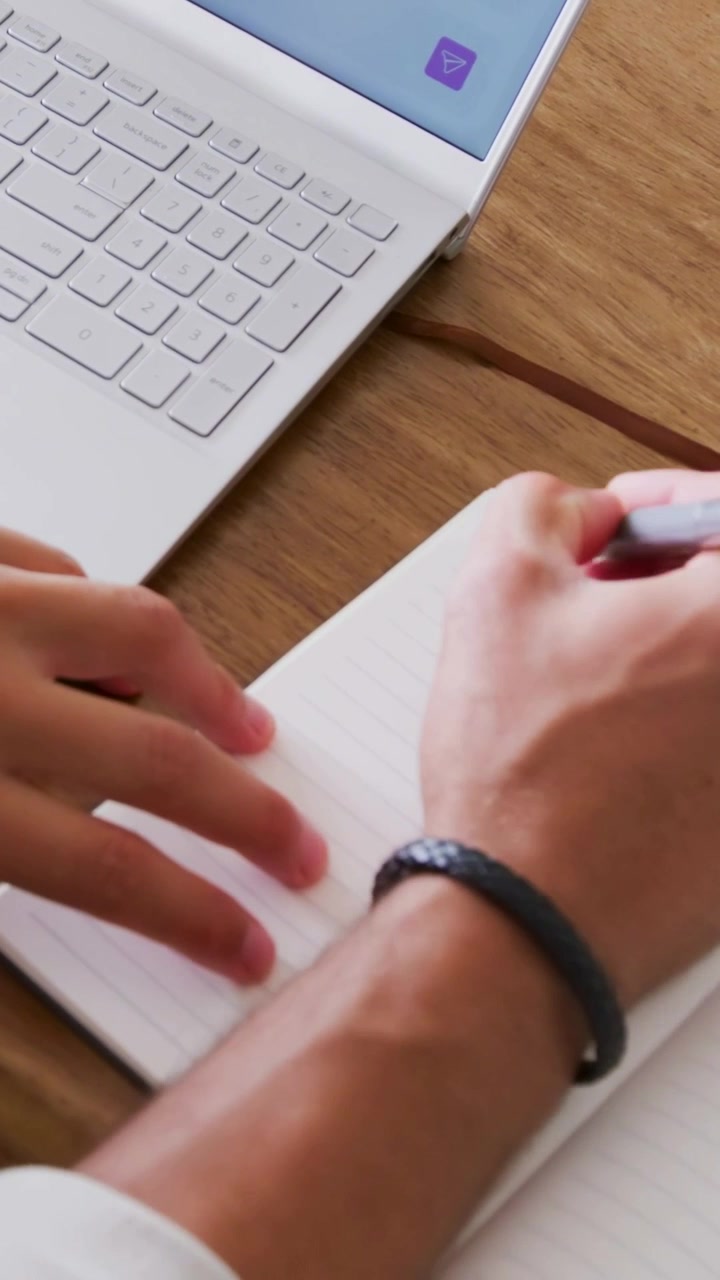 Vertical video: Noticing laptop icon, right hand writing in notebook at desk with pen and bracelet