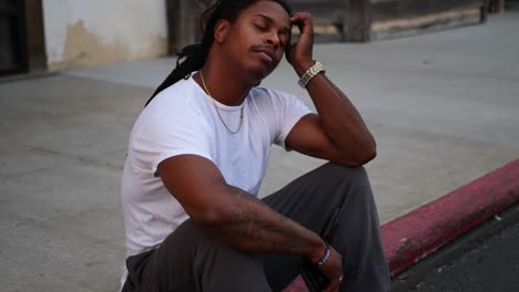 African-American-man-in-despair-sitting-on-curb-down-on-his-luck-and-sad,-hands-on-his-face,-and-eventually-looking-into-camera-after-it-pans-in