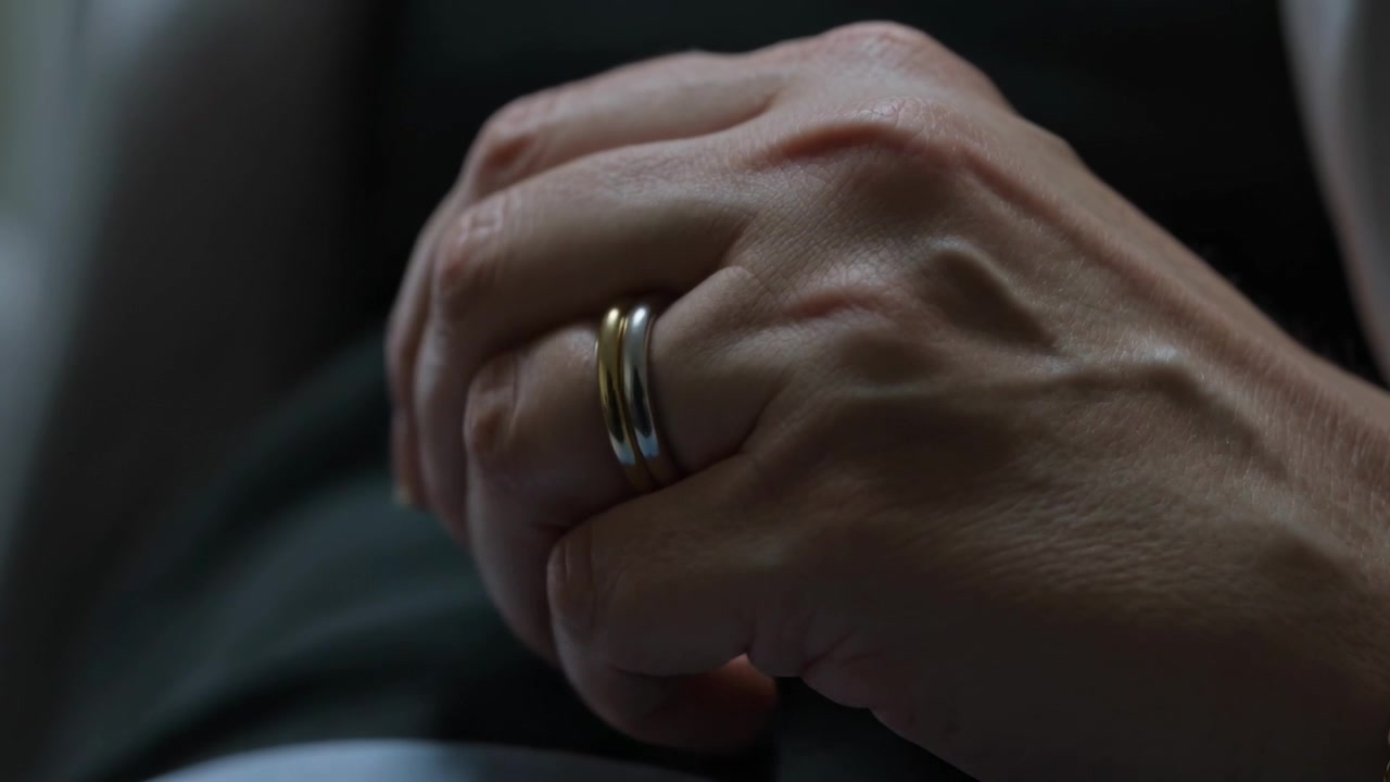 Mani di un adulto che si muovono cercando conforto in grembo su una sedia, mostrando un anello bicolore e un braccialetto d'oro.