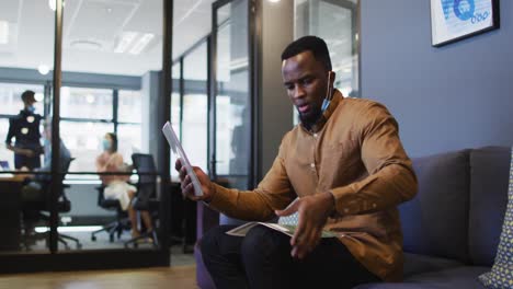 African-american-man-with-lowered-face-mask-having-video-chat-on-digital-tablet-at-modern-office