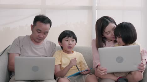 happy family, asian two young son with mom and dad sitting on sofa together. while using laptop computer browsing online internet watching cartoon, social media enjoy during holiday at home.