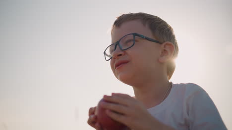 Close-up shot of a young boy wearing a white shirt and glasses, laughing as he takes a bite of a red apple in a sunlit field. The image captures a moment of pure joy and connection with nature