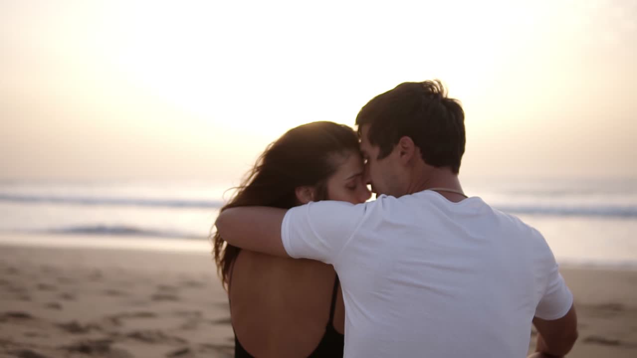 Rare view footage. Perfect scene of a loving, beautiful couple sitting on sand embraced kissing in front the ocean and mild sunset. Perfect place to be together