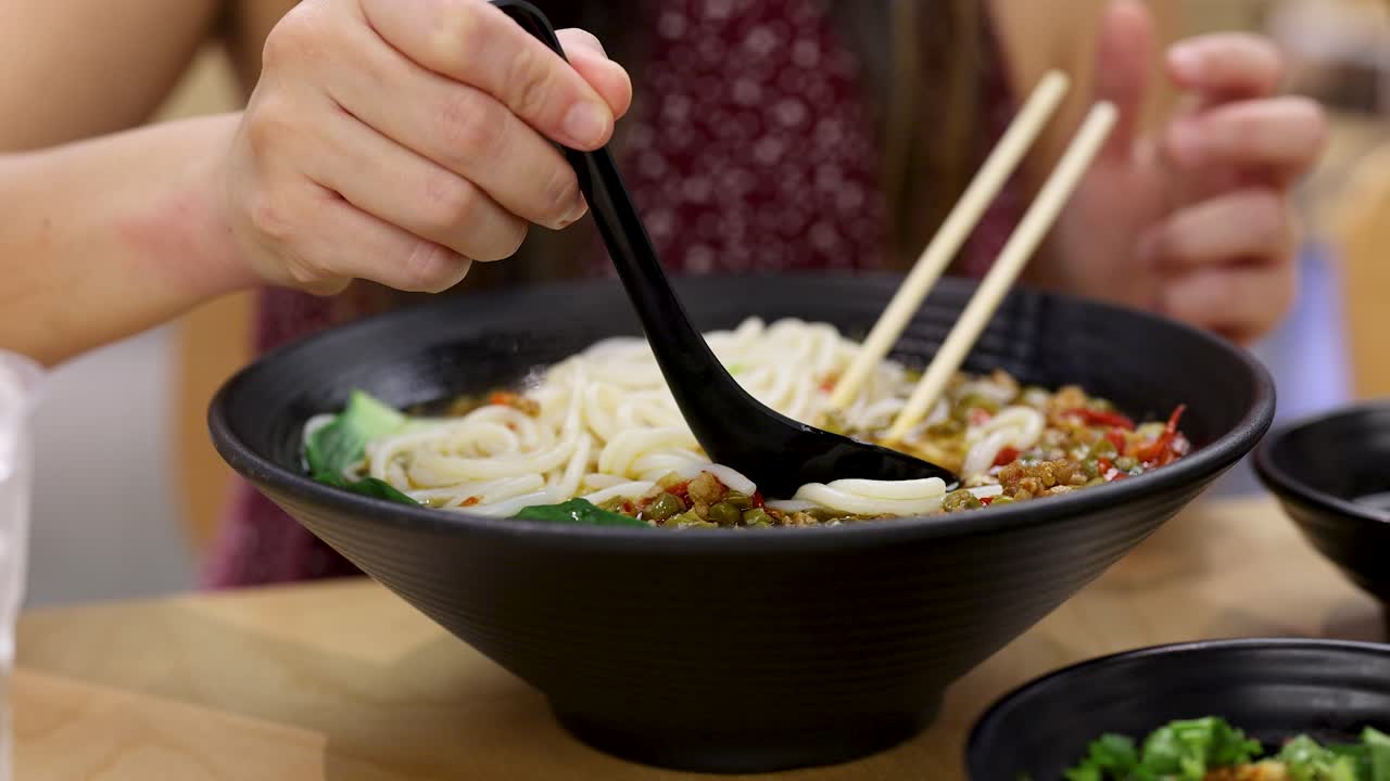 Female pours hot broth over spicy noodles in black bowl, bright indoor restaurant lighting, close-up